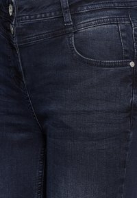 Jean en denim foncé avec un design classique à cinq poches, une texture lisse, un léger délavage et des boutons métalliques à la taille.