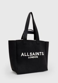 Sac cabas en toile noire avec deux poignées et texte blanc "ALLSAINTS LONDON" sur le devant, posé devant un fond clair uni.