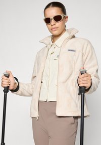 Chaqueta de forro polar crema con un patrón en relieve, cuello alto y cierre de cremallera. Camisa beige debajo, pantalones marrones y bastones en las manos. Gafas de sol con tintes marrones.