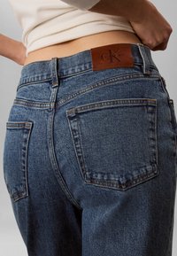 Άτομο που φοράει μπλε τζιν Calvin Klein και λευκό τοπ, δείχνοντας τις πίσω τσέπες και το δερμάτινο λογότυπο στη μέση.