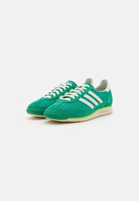 Zapatillas de ante verde con tres rayas blancas, punta redondeada, diseño plano con cordones y suela de goma texturizada.