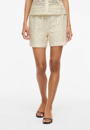 Naine, kes kannab beežikat pitsimustriga lühikesi pükse ja sobivat topi, seisab paljaste jalgadega ja kannab musti avatud varvastega sandaale.