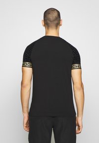 Camiseta negra de manga corta con un patrón texturizado negro y una banda de acento dorada cerca del hombro. Tejido suave, ajuste casual.