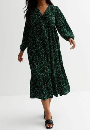 Lange groene jurk met bloemenprint, geplooide halslijn, pofmouwen en een gerimpelde zoom. Gecombineerd met zwarte sandalen met bandjes. Soepele, lichtgewicht stof.