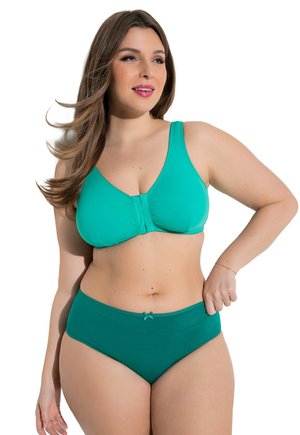 Kvinde med langt brunt hår iført en teal frontlukket bh og matchende højtaljede trusser mod en hvid baggrund, smilende let til venstre.