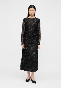 Μαύρο φόρεμα με μακριά μανίκια από διαφανές ύφασμα και περίπλοκο σχέδιο με παγιέτες σε floral μοτίβο, με λεπτομέρειες πτυχώσεων στο μπροστινό μέρος και ίσια σιλουέτα.