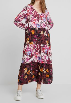 Femme portant une longue robe à fleurs avec des fleurs roses, violettes et orange, associée à des baskets blanches, debout devant un fond uni.