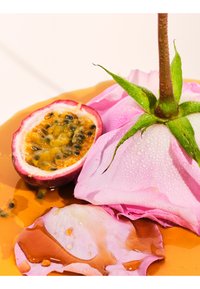 Gesneden passievrucht naast een roze roos met dauwdruppels, op een glanzend oppervlak met oranje vloeistof en verspreide rozenblaadjes.