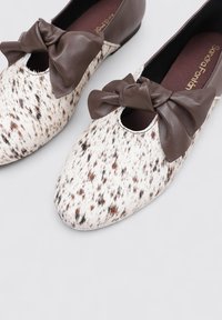 Deux chaussures plates présentant un matériau tacheté de blanc et de brun, avec un accent en cuir brun en forme de nœud et un design à bout rond.