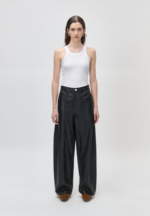 Frau mit welligem Haar trägt weißes ärmelloses Tanktop, schwarze weite Lederhose und braune Schuhe, steht vor einfachem hellem Hintergrund.