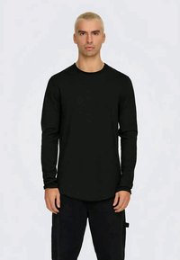 Camisola preta de manga longa feita de um tecido suave, com um decote redondo e uma bainha curva, usada com calças escuras.