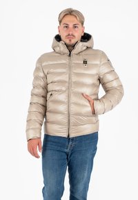Jonge man in een beige pufferjack met capuchon en blauwe jeans, staande met één hand in de zak van zijn jack tegen een witte achtergrond.
