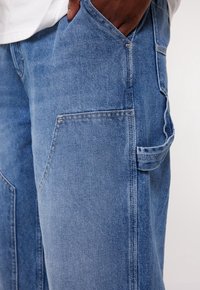 Hand delvis i framfickan på blå denim snickarbyxor med verktygslik på ljus bakgrund.