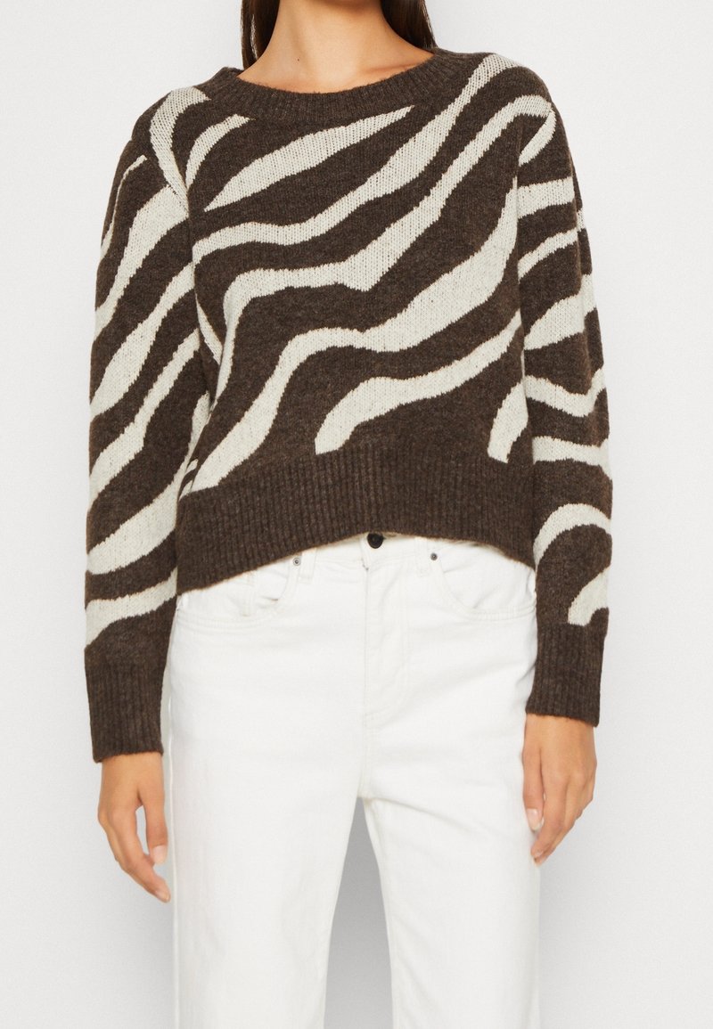 Woman wearing a brown and white zebra-striped sweater paired with high-waisted white pants against a plain background.