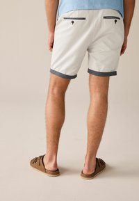 Fehér rövidnadrág sötétkék szegéllyel, két hátzseb gombos zárással, barna slip-on cipőkkel viselve. Sima anyagtextúra.