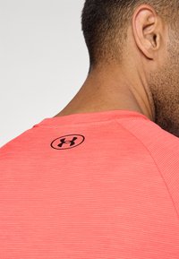 Punainen urheilupaita, jossa on musta logo takakauluksessa. Mallissa on pyöreä pääntie ja sileä, istuva muotoilu.