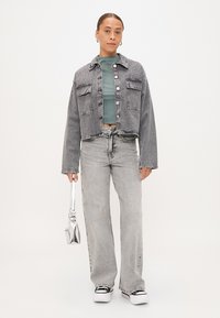 Chaqueta de mezclilla gris cortada con dos bolsillos en el pecho y botones plateados, llevada sobre una blusa verde y combinada con jeans acampanados gris claro y zapatillas blancas.
