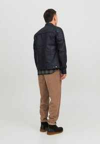 Giacca nera in finta pelle con spalle texturizzate, indossata sopra una camicia a quadri; abbinata a pantaloni beige in corda e stivali neri.