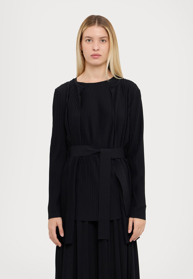 Schwarzes, plissiertes Langarmoberteil mit Rundhalsausschnitt und Bindegürtel in der Taille, aus einem strukturierten Stoff gefertigt, mit vertikalen Falten.