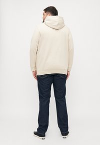 Beige Kapuzenpullover mit weicher Textur, kombiniert mit dunkelblauen Jeans und schwarzen Sneakers. Ansicht von hinten, die den Winkel der Kapuze zeigt.