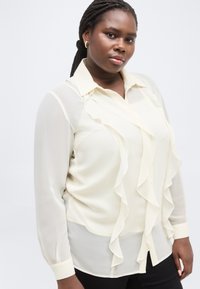 Αυτοπεποίθητη γυναίκα με πλεξούδες φορά διαφανή, εκρού μπλούζα με βολάν, συνδυασμένη με μαύρο παντελόνι, πάνω σε απλό φόντο.