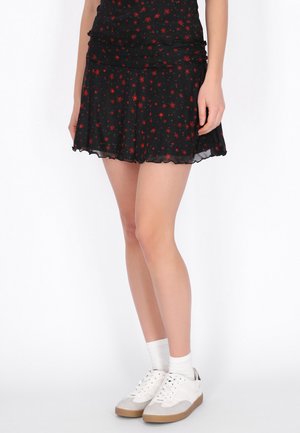 Frau trägt einen schwarzen Minirock mit rotem Blumenmuster, weiße Knöchelsocken und weiße Turnschuhe, steht vor einem einfachen weißen Hintergrund.