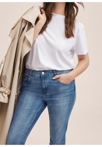 Femme portant un t-shirt blanc et un jean bleu, une main dans la poche, tenant un trench-beige sur l'épaule, sur un fond neutre.