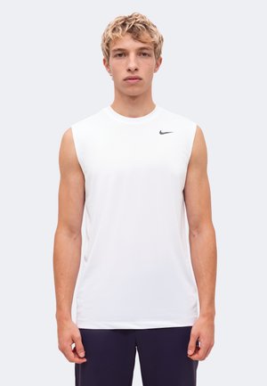 Млад мъж позира с бяла тениска Nike без ръкави и тъмни къси панталони на изчистен светъл фон.