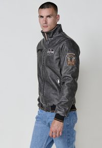 Chaqueta bomber de cuero sintético gris con cuello alto, cierre de cremallera, parches bordados en la manga y puños y dobladillo acanalados.