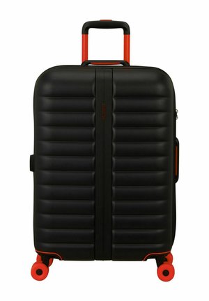 Maleta rígida negra con relieves horizontales, asa telescópica roja y cuatro ruedas giratorias rojas.