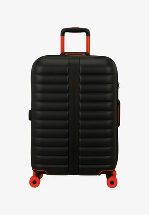 Valise rigide noire avec des rainures horizontales, une poignée télescopique rouge et quatre roues rouges pivotantes.