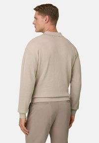 Homme de profil portant un pull beige en maille côtelée et un pantalon beige assorti sur un fond uni.