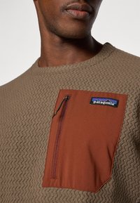Brauner strukturierter Sweatshirt mit einer kontrastierenden braunen Reißverschlusstasche und einem Patagonia-Logo-Patch auf der Brust, mit einem runden Halsausschnitt.