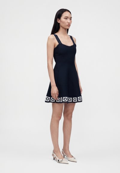 Femme debout portant une robe noire sans manches avec de larges bretelles et une bordure florale blanche à l'ourlet, associée à des chaussures beiges à talons bas.