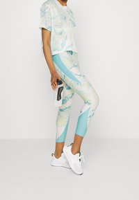 Lekka siateczkowa bluzka crop top z pastelowym nadrukiem liści. Legginsy z nadrukiem mają niebiesko-białą geometryczną wzór. Białe buty sportowe.