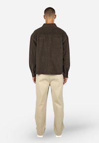 Barna kordbársony ingdzseki laza szabású, egy mellzsebbel, gombos mandzsettával, világosbarna laza szabású nadrággal és fehér cipőkkel párosítva.