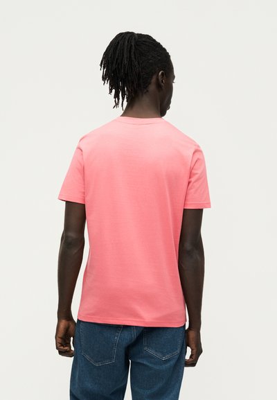 Homme à la peau foncée et aux dreadlocks, portant un t-shirt rose uni à manches courtes et un jean bleu, debout de dos sur un fond uni.