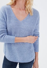 Jasnoniebieski, miękki sweter z dzianiny z dekoltem w serek, długimi rękawami i prążkowanymi mankietami. Ma zaokrąglony dół oraz lekko puszystą fakturę.