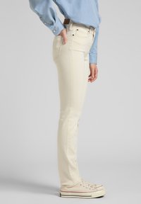 Persona vestida con una camiseta de manga larga azul claro, jeans slim color crema y zapatillas de lona crema, de pie con una mano en el bolsillo trasero contra un fondo gris.