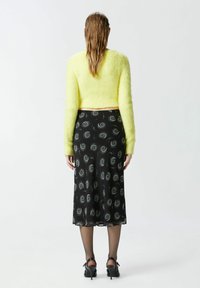 Žuti plišasti skratjeni džemper u kombinaciji s crnom midi suknjom s bijelim uzorcima spirala, upotpunjen prozirnim crnim tajicama i crnim potpeticama.