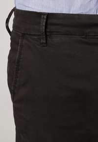 Schwarze Baumwollhose mit flachem Bund, sichtbaren Nähten und Gürtelschlaufen. Der Stoff ist glatt mit matter Oberfläche.