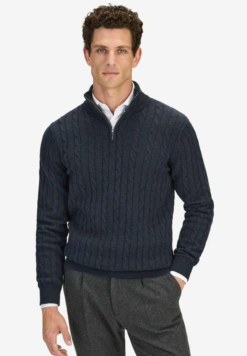 Marinblå kabelstickad tröja med halvdragkedja och ribbade ärmslut, tillverkad av ett mjukt material, bärs över en vit skjorta med krage.