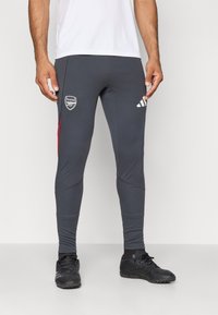 Leggings de sport gris à design ajusté, avec des accents rouges, un logo Arsenal et une marque Adidas sur la hanche. Fabriqués à partir d'un matériau extensible.