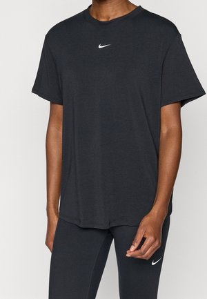 Personne portant un t-shirt noir Nike et des leggings noirs, tous deux arborant le logo blanc Nike swoosh, debout devant un fond uni.