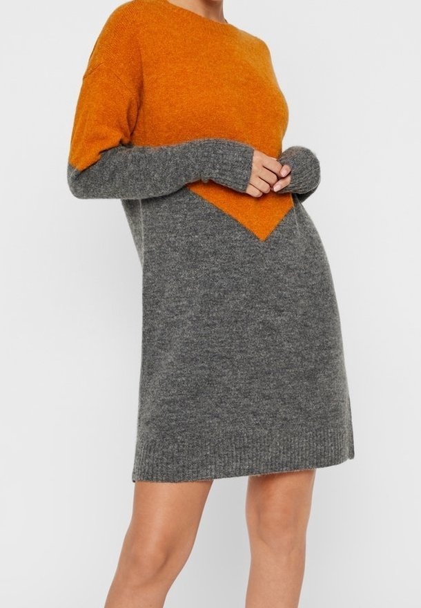Femme portant une robe tricotée à manches longues, haut orange et bas gris, debout, les mains légèrement jointes devant.