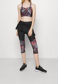 Crop top sportivo nero con motivi geometrici rossi e grigi, abbinato a leggings neri a 3/4 con accenti coordinati. Sneakers nere completano il look.