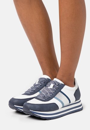 Chaussures de sport blanches et bleu marine avec un tissu texturé, une semelle rayée et un laçage unique. Les accents comprennent des détails argentés et bleus sur les côtés.