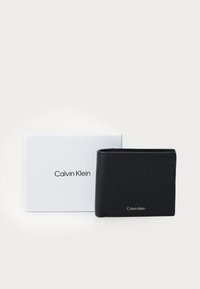 Juodas, tekstūruotas odinis piniginė su sulankstomu dizainu, eksponuojama šalia baltos dėžutės su juodu "Calvin Klein" prekių ženklu.