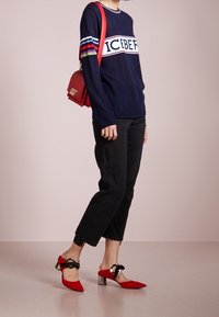 Granatowy sweter w paski z napisem „ICEBERG”, czarne krótkie spodnie oraz czerwone szpilki z ostrym czubkiem, srebrnymi blokowymi obcasami i kokardkami. Czerwona torebka.