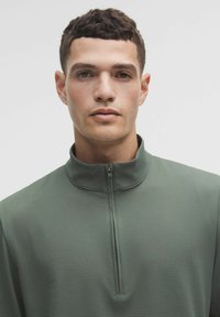 Zelený zipsový pulover vyrobený z texturovanej látky s jemným bodkovaným vzorom. Golier sa zdvíha a má predný zips.